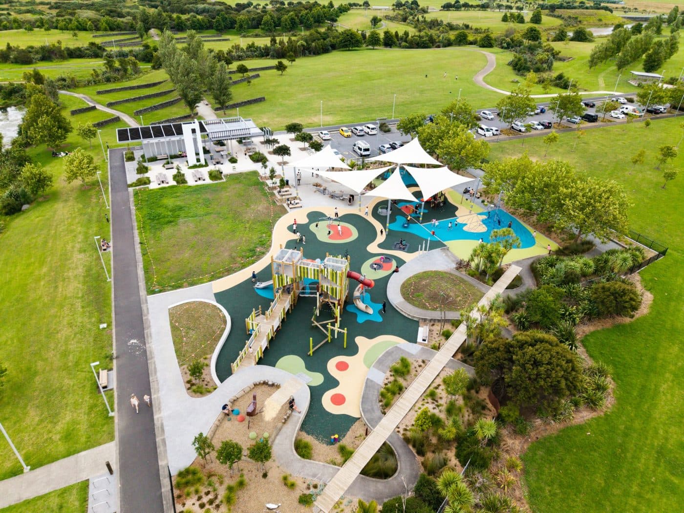 Aerial view of a modern playground with white shade sails and colourful surfacing, next to open sports fields